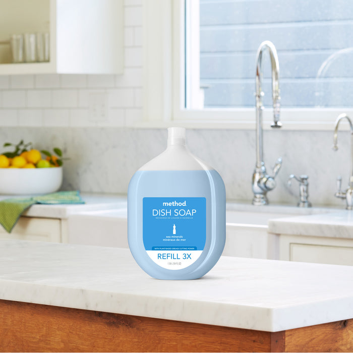 A large Method Dish Soap Refill with a blue label sits on a kitchen countertop made of white marble, next to a stainless steel faucet. Sunlight streams through the window, gently lighting the room, while a fruit bowl graces the background.