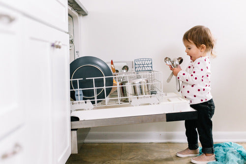 A young child stands in front of an open dishwasher, holding silverware and looking at the dishes—thanks to Seventh Generation Dish Soap Refill, parents can confidently let little helpers join the clean-up.