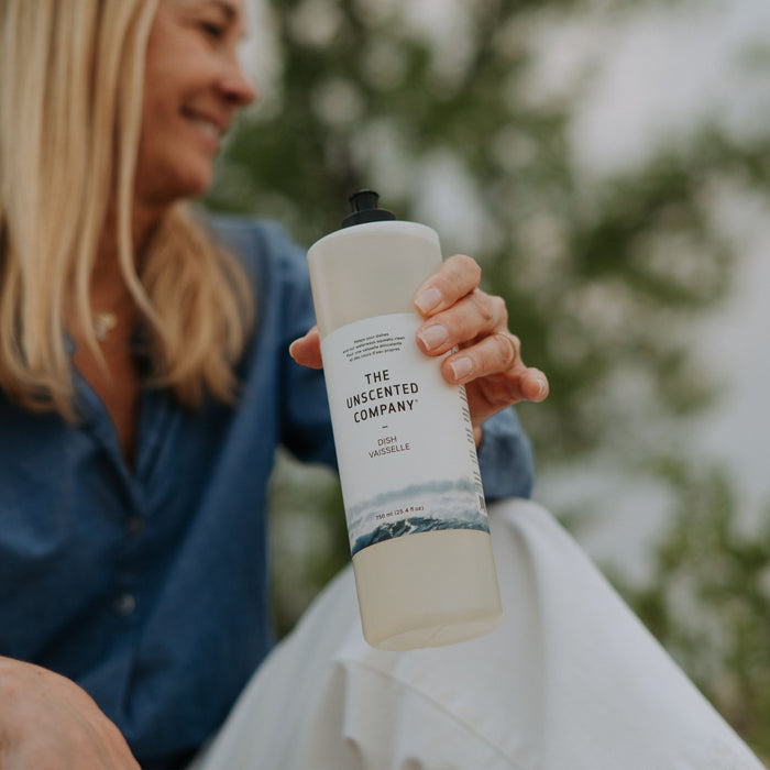 A smiling woman with long blonde hair holds a bottle of The Unscented Company Dish Soap outdoors, with greenery blurred in the background.