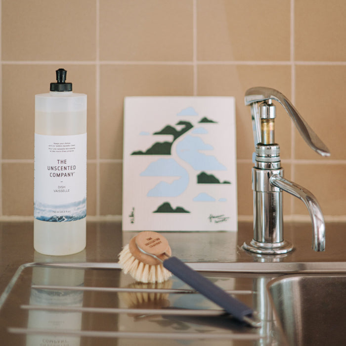 A bottle of The Unscented Company Dish Soap, a dish brush, and a scrubber sit on a kitchen counter beside a modern faucet and a decorative card with mountain and river art, all set against a tiled wall.