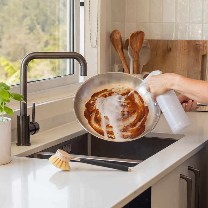 At the kitchen sink, a person uses Cleanerys Dish Spray Starter Kit to clean a greasy frying pan, with a spray bottle in hand and a nearby scrubbing brush. Wooden utensils rest against the bright window, offering an ideal eco-friendly dishwashing scene.