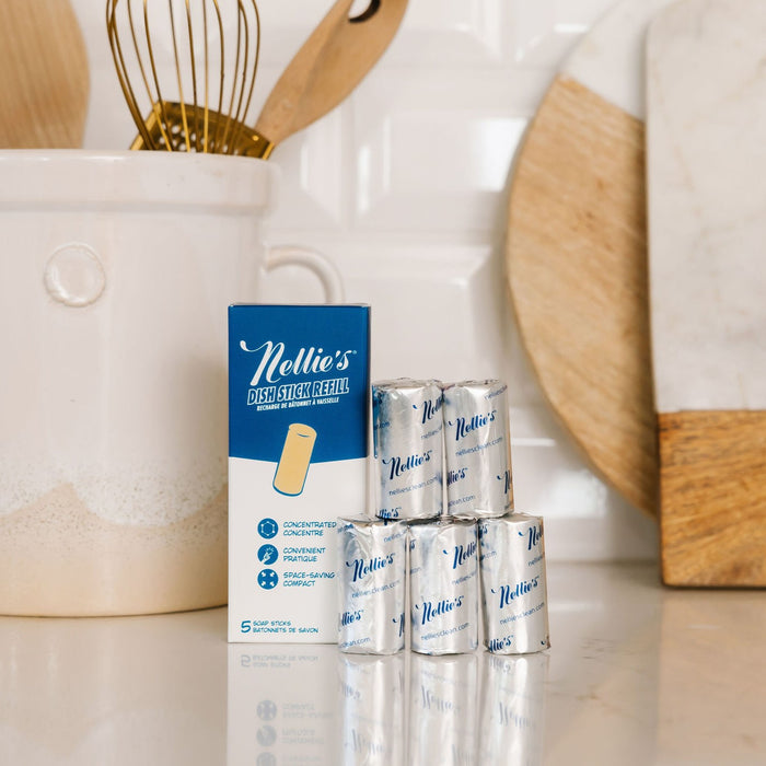 A kitchen counter features Nellies Dish Stick Refill Soap box with silver refills stacked nearby. In the background, a white ceramic utensil holder with wooden spoons and a cutting board completes this eco-friendly cleaning setup.