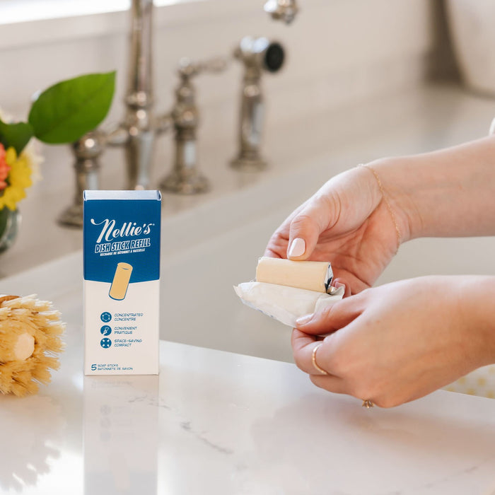 Close-up of hands unwrapping a Nellies Dish Stick Refill Soap by a faucet. A box labeled Dish Stick Refill and a scrub brush are on the countertop, set in a bright, clean kitchen with sunlight.