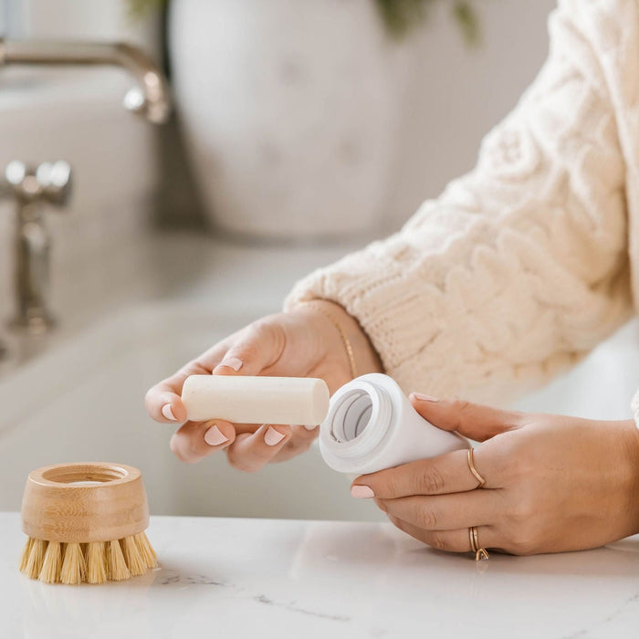 In a bright kitchen, someone in a cream sweater uses Nellies Dish Stick Refill Soap to clean, holding the wooden-topped round dish brush. The contrasting white countertop highlights the faucet and blurred background elements as they remove a white refillable container.