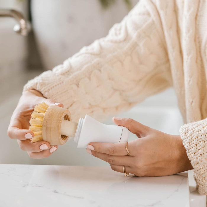 A person in a beige cable-knit sweater is using Nellies Dish Stick Refill Soap, which has a wooden handle, over the countertop as they assemble or disassemble the bristle brush for their eco-friendly cleaning routine.