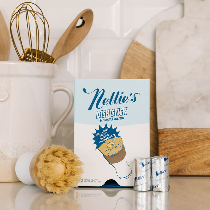 On the kitchen counter, a box of Nellies Dish Stick takes center stage, surrounded by utensils in a ceramic holder. Nearby sit a scrub brush and foil-wrapped items on an eco-friendly surface, with a marble cutting board in the background.