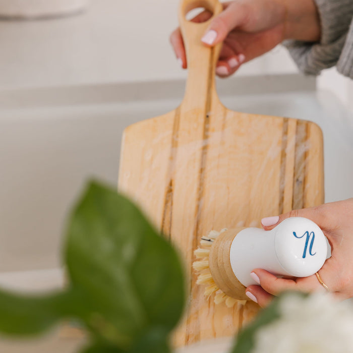 In a white sink, someone uses a Nellies Dish Stick to scrub a wooden cutting board, promoting green cleaning habits. The brush features a white handle with a blue n logo. A blurred green plant in the foreground adds to the eco-friendly ambiance.