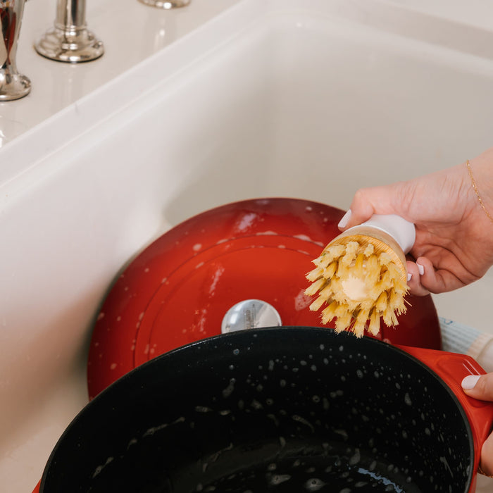 Someone uses a Dish Stick by Nellies over a sparkling white sink, scrubbing a large red pot with a black interior, while water splashes around, highlighting an eco-friendly cleaning method.