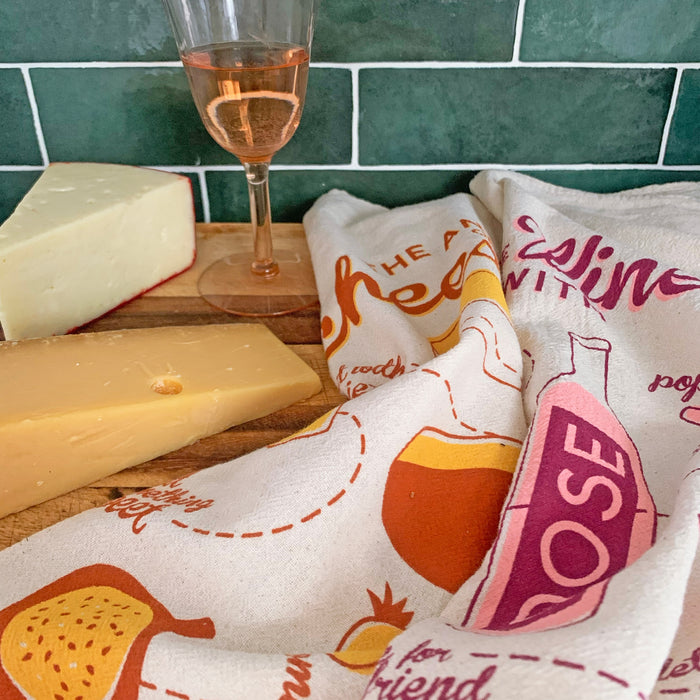 A glass of rosé, two cheese blocks, and The Neighborgoods Dish Towel Set with wine and cheese designs are displayed on a wood surface against a green tile backdrop.