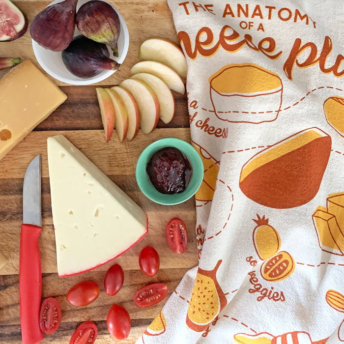 A wooden board with cheese, figs, apples, tomatoes, and jam sits beside a red-handled knife and The Neighborgoods Dish Towel Set featuring a colorful Anatomy of a Cheese Plate design.