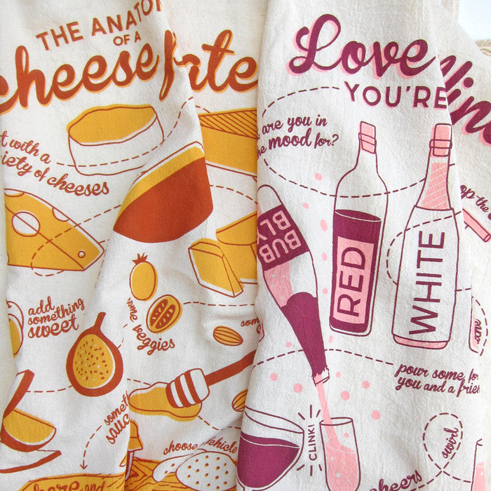The Neighborgoods Dish Towel Set includes two illustrated cotton towels—one with orange and yellow cheese designs, the other with pink and purple wine bottles, glasses, and playful text—perfect for brightening your kitchen collection.