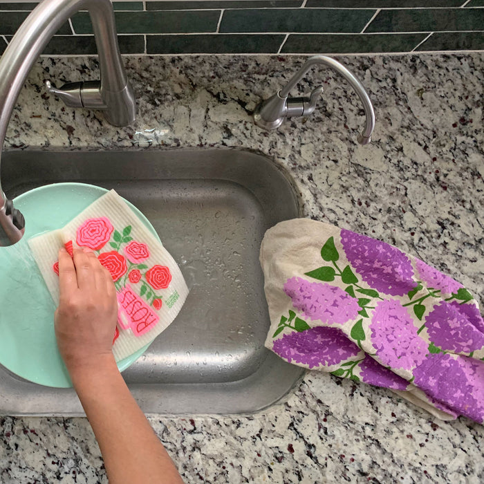 Someone washes a light blue plate in the kitchen sink using a floral-patterned cloth. Adjacent to the sink, an absorbent dish towel by The Neighborgoods rests on the granite countertop, matching the beige towel with purple flower designs.