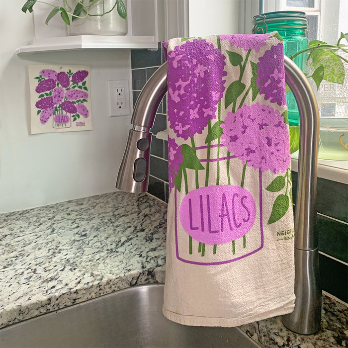 A kitchen sink with a curved faucet has The Neighborgoods unbleached cotton Dish Towel, known for its super absorbency, draped over it. The towel displays purple lilacs in a jar with Lilacs text. Similar artwork decorates the wall, and potted plants are on the windowsill.