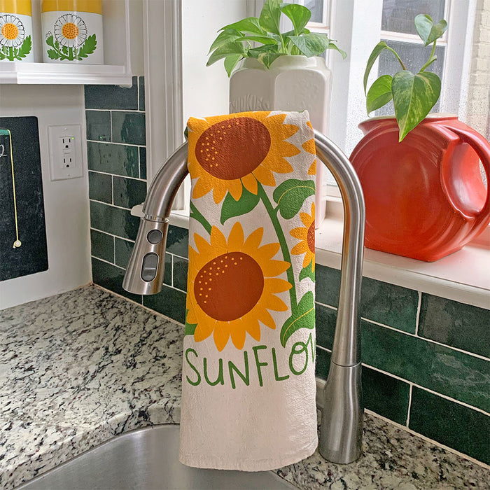 A kitchen faucet showcases The Neighborgoods absorbent Dish Towel featuring sunflower illustrations and the word sunflower. A red teapot and a green plant enhance the windowsill against a green tiled backsplash and granite countertop.