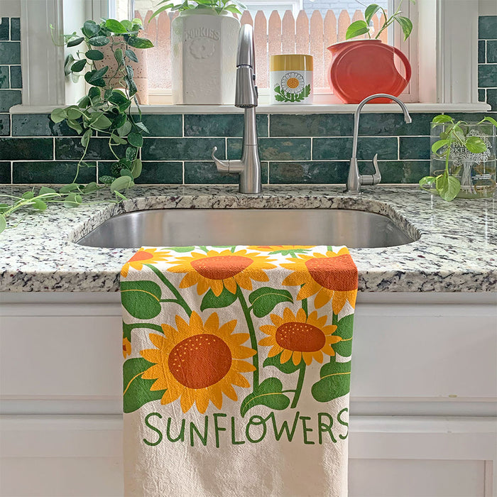 A kitchen sink featuring The Neighborgoods Dish Towel made of unbleached cotton, adorned with bright yellow sunflowers, green leaves, and the word SUNFLOWERS. The backdrop showcases green tile, potted plants, and other kitchen essentials.