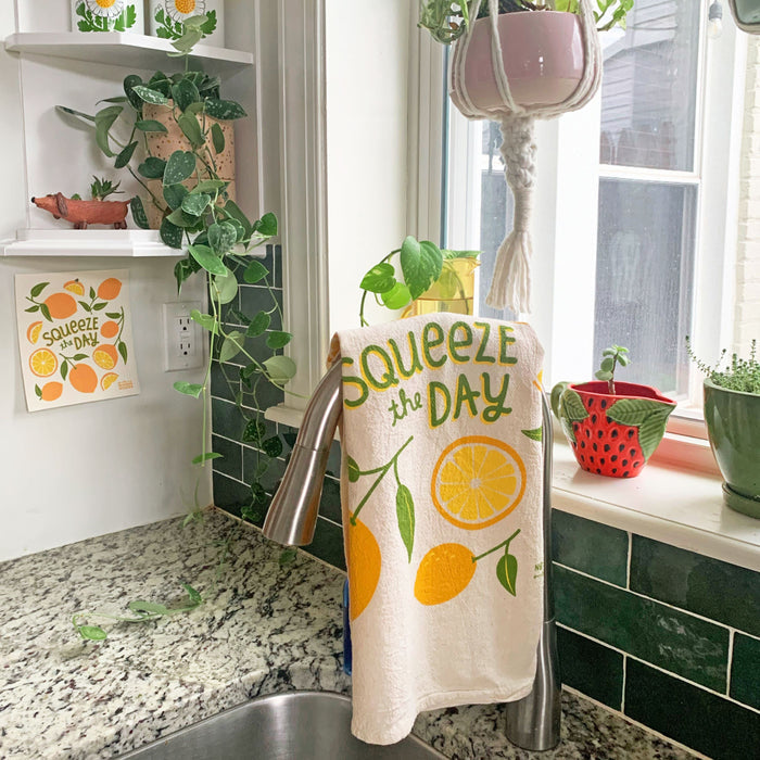 A kitchen sink area showcases The Neighborgoods absorbent dish towel on the faucet, featuring a Squeeze the Day lemon theme. Greenery and decor items abound, including a small potted plant on the windowsill and coordinating lemon-themed wall art.