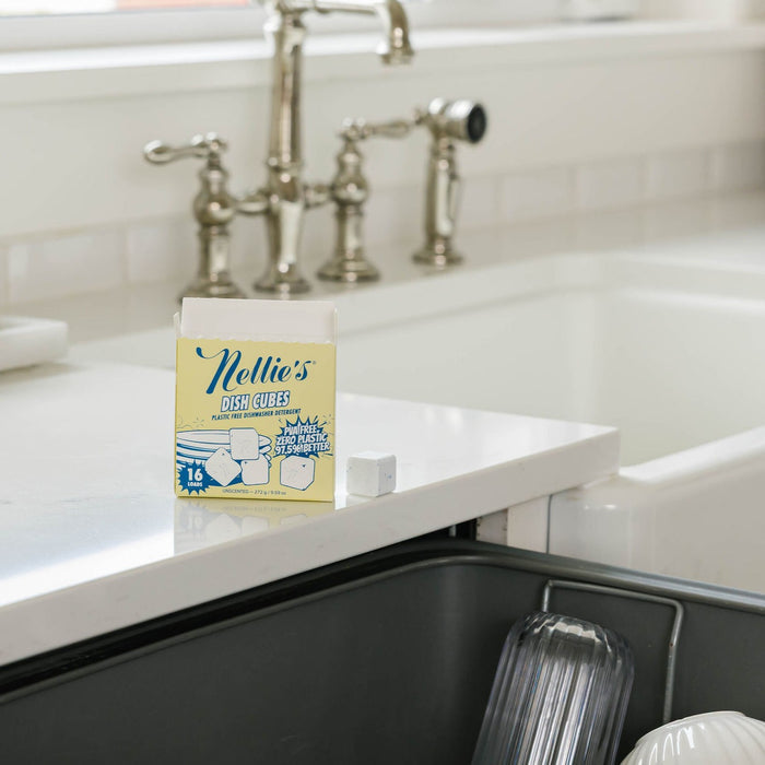 A box of Nellie's Dish Cubes, 16 Load, rests on a white kitchen counter by a farmhouse sink and open dishwasher. One eco-friendly dish cube is placed next to the box.