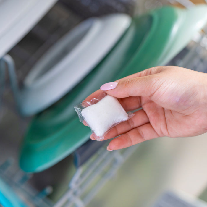 A hand places a Seventh Generation Dishwasher Detergent Pack into an open dishwasher. In the background, the enzyme-rich formula effectively cleans white and teal dishes neatly arranged on the racks.