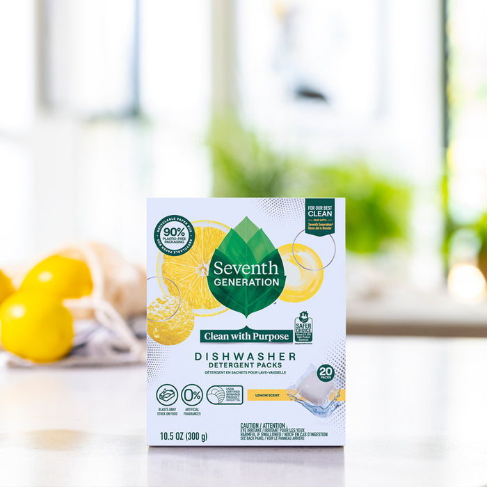 A box of Seventh Generation Dishwasher Detergent Packs with a lemon scent rests on a kitchen counter. The packaging highlights an enzyme-rich, chlorine-free formula, promoting eco-friendliness. In the background, blurred lemons and folded towels enhance the scene.
