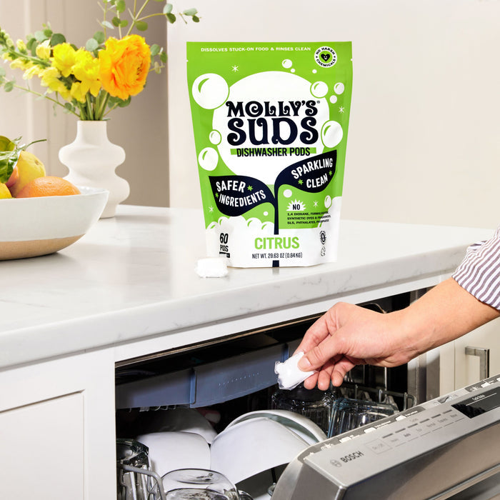 A person puts a Molly’s Suds Dishwasher Detergent Pod into a loaded dishwasher. A bag of Molly’s Suds Dishwasher Detergent Pods sits on the counter with flowers and fruit in the background.