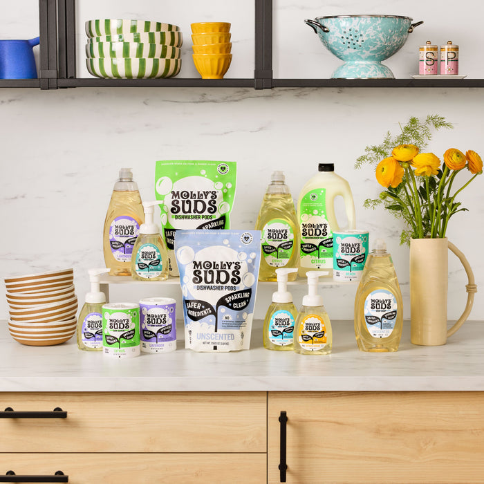 On the kitchen countertop, Molly’s Suds Dishwasher Detergent Pods are displayed among other cleaning products. Behind them, bowls and dishes are stacked on shelves, with a vase of yellow flowers on the right.