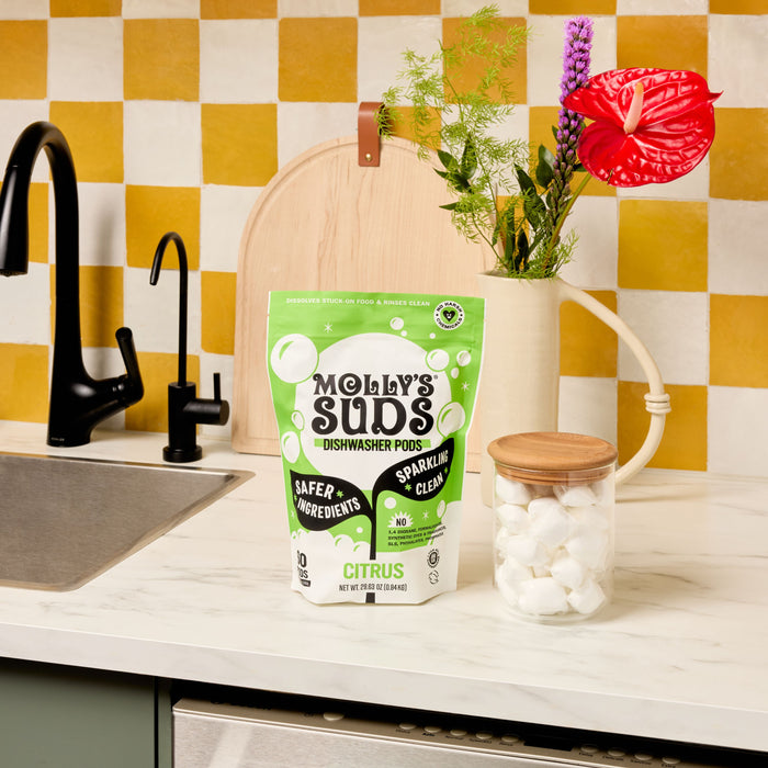 A bag of Molly’s Suds Dishwasher Detergent Pods sits on a white kitchen counter beside a glass jar of pods, with a nearby sink and decorative flowers. A yellow and white checkerboard tile wall adds brightness to the scene.