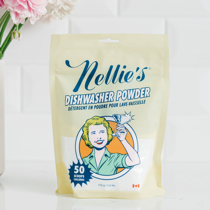 A pouch of Nellie's Dishwasher Powder sits on a white counter. The pale yellow bag with retro artwork boasts 50 scoops, is phosphate-free and septic safe. Pink flowers decorate the background for a cheerful touch.