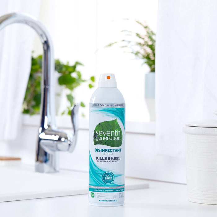 A Seventh Generation Disinfectant Spray is displayed on a kitchen counter next to a chrome faucet, highlighting its ability to kill 99.99% of bacteria and viruses on nonporous surfaces, with blurred greenery and a white container in the background.