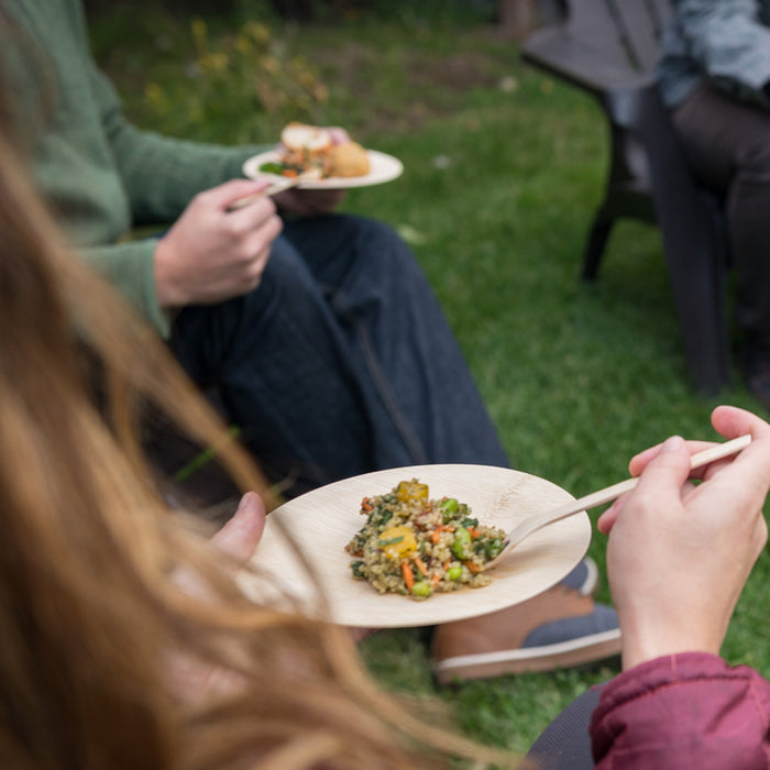 Two casually dressed people enjoy a meal of quinoa salad outdoors on the grass, each holding a Bambu Disposable Bamboo Plate.