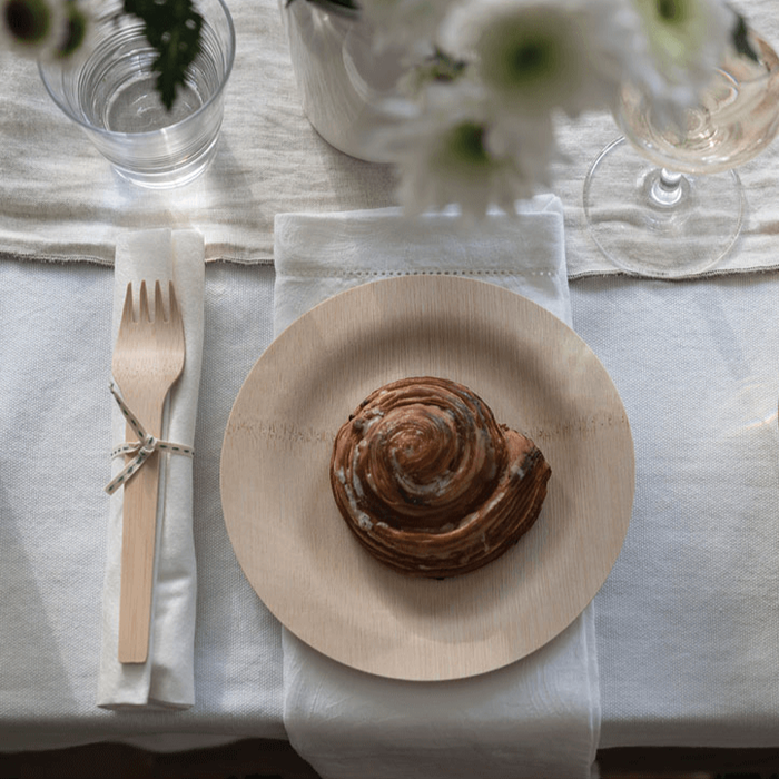 A croissant sits on a Bambu Disposable Bamboo Plate, set on a neat table with a cloth napkin and wooden cutlery tied with string, accompanied by a glass of water, a wine glass, and flowers in the foreground.
