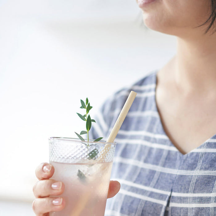 Sipping a light pink drink from a textured glass with green herb garnish, they use a beige Disposable Bamboo Straw by Bambu while wearing a blue and white checkered top against a softly blurred background.