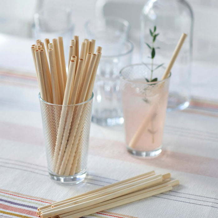 A glass of sturdy beige Bambu Disposable Bamboo Straws is on a table with more straws beside it. Nearby, a herb-garnished drink in another glass sits against a striped tablecloth backdrop, complemented by clear glasses, celebrating plastic alternatives.
