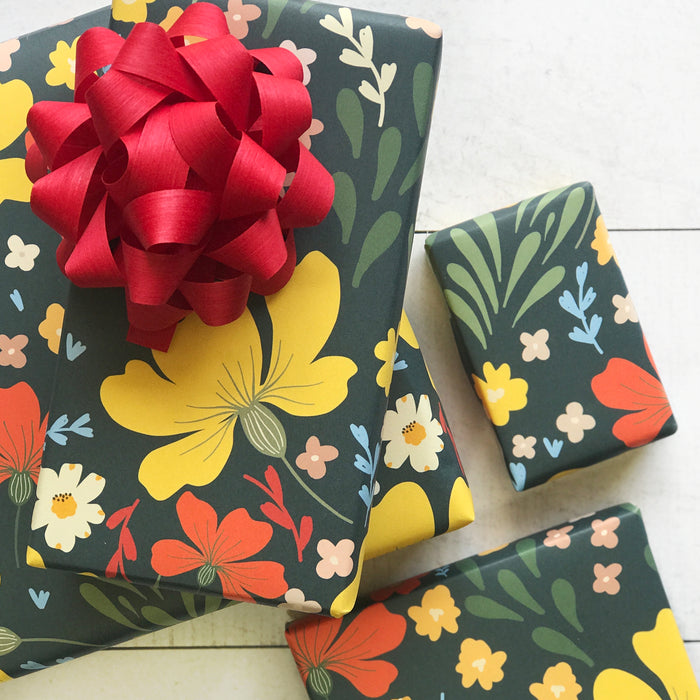 Gifts are wrapped in Dissolvable Gift Wrap by Waterleaf Paper Company, featuring yellow, red, and white flowers on a dark background. A large red bow adorns one gift. Made with eco-friendly techniques, they are beautifully arranged on a light wooden surface.