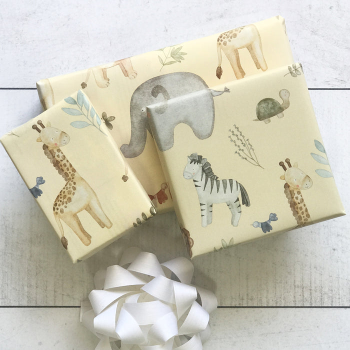 Three presents wrapped in Waterleaf Paper Companys Dissolvable Gift Wrap, featuring giraffes, an elephant, a zebra, and a turtle on animal-themed paper, sit on a white surface with a white ribbon bow in front. This eco-friendly packaging offers an innovative touch to gift-giving.