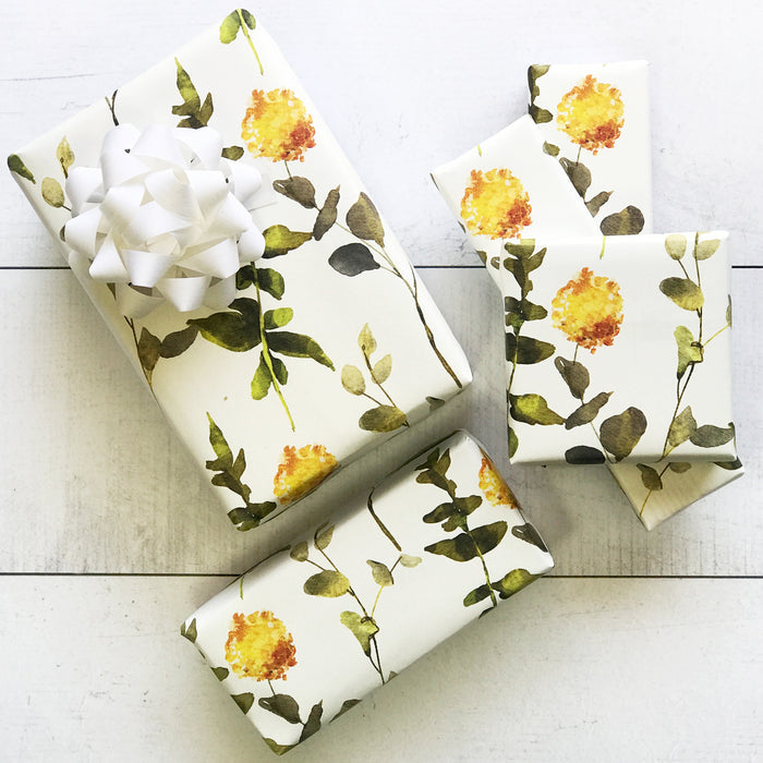 Three gifts wrapped in Waterleaf Paper Companys Dissolvable Gift Wrap, featuring a floral pattern with yellow flowers and green leaves printed using water-based ink, are arranged on a white wooden surface. One gift is topped with a white ribbon bow.