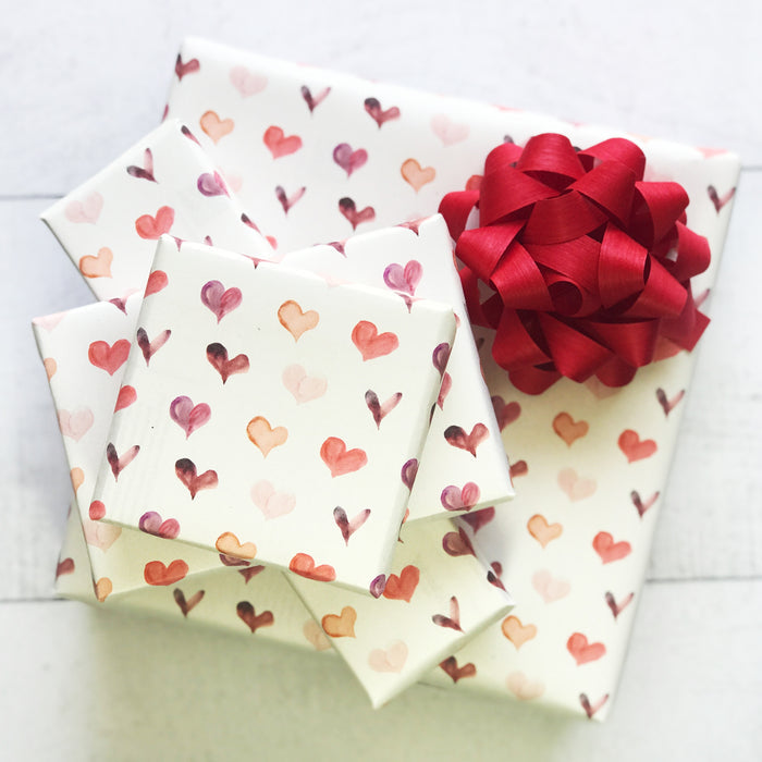 Four gift boxes of varying sizes are stacked, wrapped in Waterleaf Paper Companys Dissolvable Gift Wrap featuring pink, orange, and red hearts. The largest box is topped with a large red bow, making the set both charming and eco-friendly.