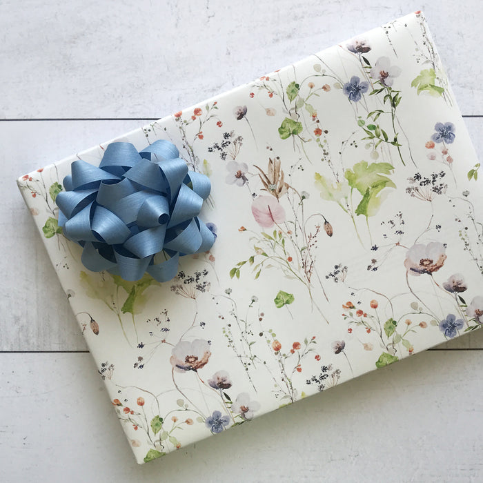 A Dissolvable Gift Wrap from Waterleaf Paper Company, featuring eco-friendly floral patterns in pink, blue, and green, is adorned with a large blue ribbon bow and set on a light wooden surface.