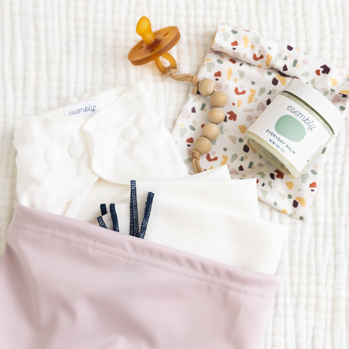 A collection of baby essentials featuring a pacifier with a wooden clip, Ditty Bag Duo Wet Bags by Esembly Baby, and versatile white cloth diapers with navy and gray accents. A jar labeled everyday balm completes the set, elegantly arranged on a textured white surface.