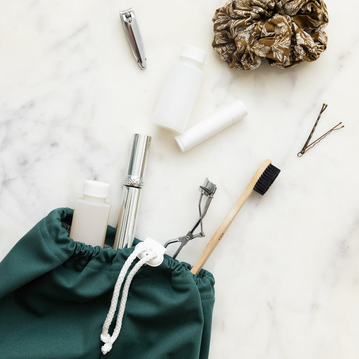 A green Ditty Bag Duo Wet Bag by Esembly Baby, crafted from recycled plastic, holds a wooden toothbrush, eyelash curler, and nail clipper on a marble surface. Nearby are bottles, lip balm, a patterned scrunchie, and bobby pins.