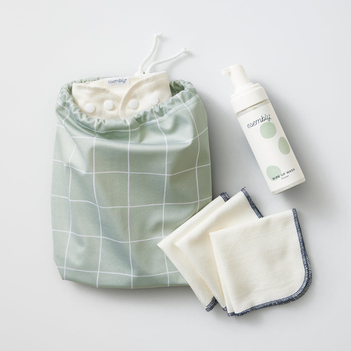The Ditty Bag Duo Wet Bags, by Esembly Baby, showcase two white cloth wipes with a dark edge. A green grid-patterned reusable diaper with recycled plastic and white snaps sits next to foaming baby bum wash. The diaper features an open design and drawstring closure.