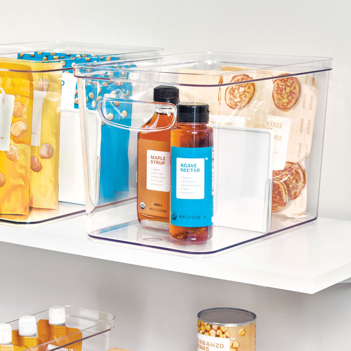 An iDesign Divided Organizer Bin with Handles, made from recycled plastic, sits on a white shelf holding maple syrup and agave nectar bottles. Nearby, similar bins neatly store packaged goods like pancake mix, all ensuring safe and efficient home organization with BPA-free construction.