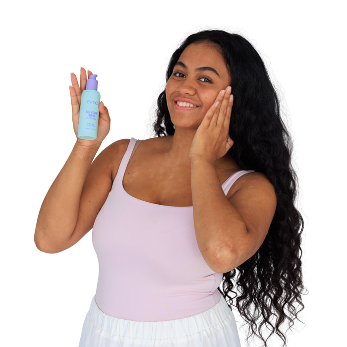 A woman with long hair smiles, holding evre.s Do Your Thing AHA & BHA Gel Cleanser for Oil Control in one hand while touching her cheek. She wears a light pink tank top and white skirt against a plain white background, showing clear skin free from blackheads and breakouts.