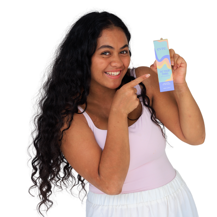 A woman with long curly hair, wearing a white tank top and pants, smiles while pointing to a colorful box labeled DO YOUR THING by evre. The pastel-colored box contains the Do Your Thing AHA & BHA Gel Cleanser for Oil Control, designed for gentle exfoliation.