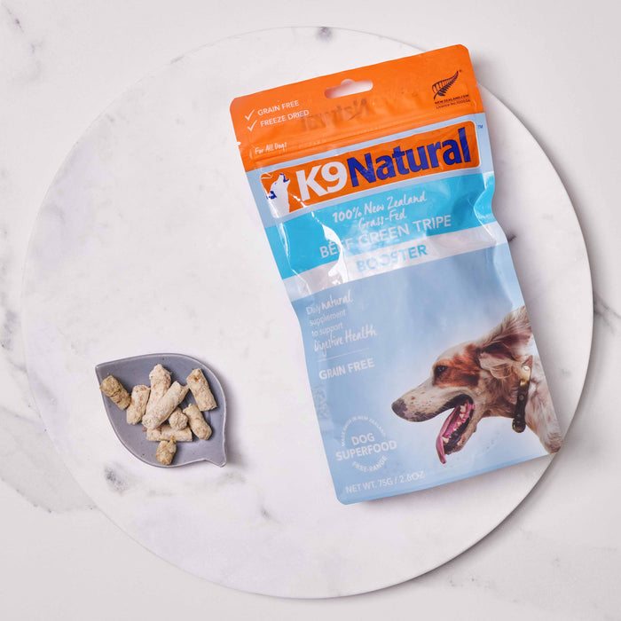 A package of K9 Natural Grain-Free Freeze-Dried Dog Food Supplement Booster, premium dog food from New Zealand, sits on a round white marble surface beside a small gray dish with pebble-like treats from the Freeze-Dried Boosters collection.