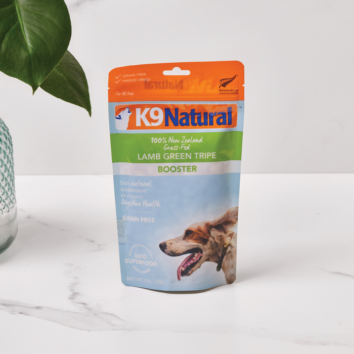 On a marble surface rests a package of K9 Naturals Grain-Free Freeze-Dried Dog Food Supplement Booster featuring New Zealand Lamb Tripe. The pouch has a dog image and promotes its Grain Free, Dog Superfood benefits, with a green plant partially visible on the left.