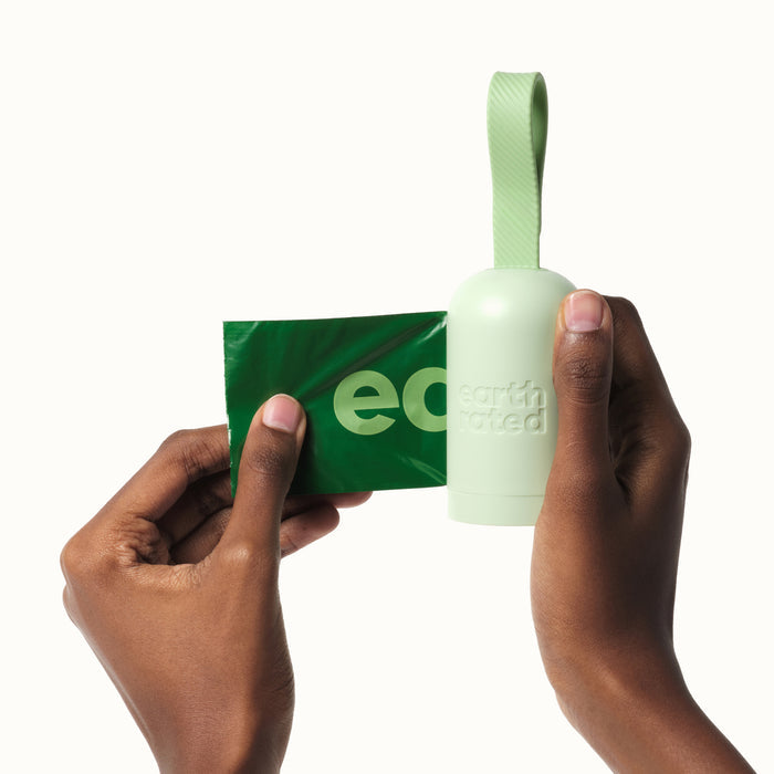 Hands holding the Earth Rated Dog Leash Dispenser, crafted from recycled plastic with a textured strap, while a green waste bag is pulled out. The dispenser and bag duo guarantee sustainability and reliability, with Earth Rated embossed on its surface.
