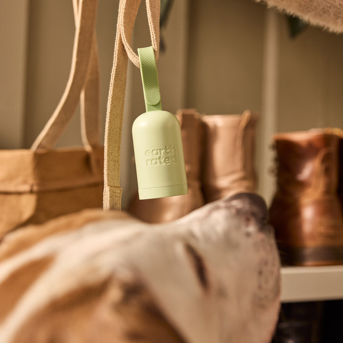 An Earth Rated light green Dog Leash Dispenser, made from recycled plastic, hangs from a strap. In the cozy, warmly lit background, brown boots and a dogs head contribute to the rustic atmosphere.