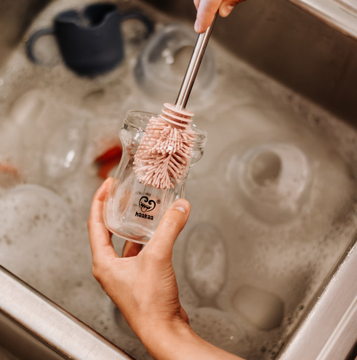 Over a soapy sink, someone cleans a glass baby bottle using Haakaas pink Double-Ended Silicone Cleaning & Bottle Brush. The bottle, featuring a heart logo and haakaa branding, lies amidst other submerged kitchen items.