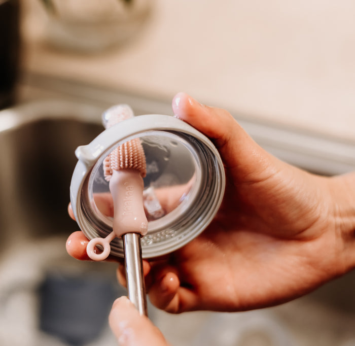 A pair of hands washes a baby bottle with Haakaas Double-Ended Silicone Cleaning & Bottle Brush, emphasizing hygiene essentials. The spotless, transparent bottle contrasts against the softly blurred sink in the background.