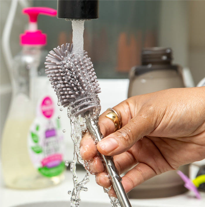 A person cleans a Haakaa Double-Ended Silicone Cleaning & Bottle Brush under running water at a sink, with a pink-pumped liquid soap bottle visible in the background, highlighting hygiene essentials as water droplets splash around.